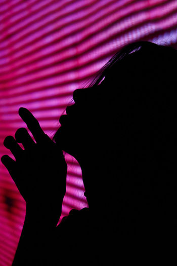 Side view silhouette of anonymous sensual female model gently touching chin near wall with glowing lights in dark studio