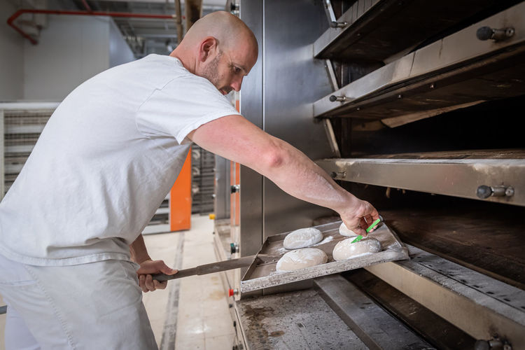 Side view baker scoring unbaked bread loaf with knife on flat shovel into electric oven while working in bakery