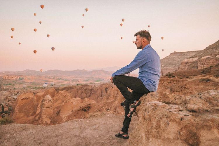 Side view of man sitting on rock with hot air balloons flying background