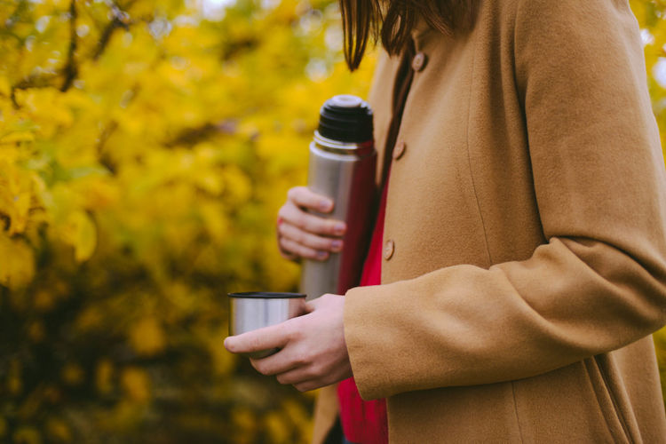 Midsection of woman holding insulated drink container while standing in forest during autumn