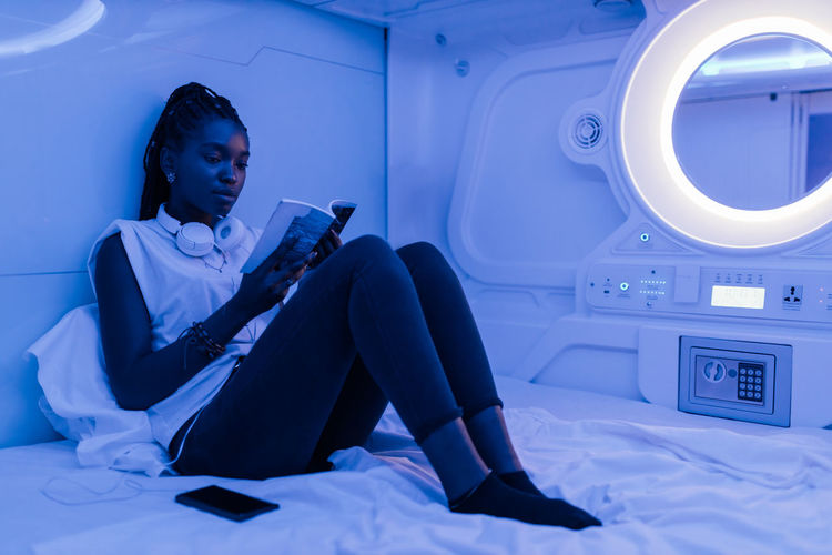 Young black female tourist sitting on bed and reading book while relaxing in room of contemporary capsule hotel