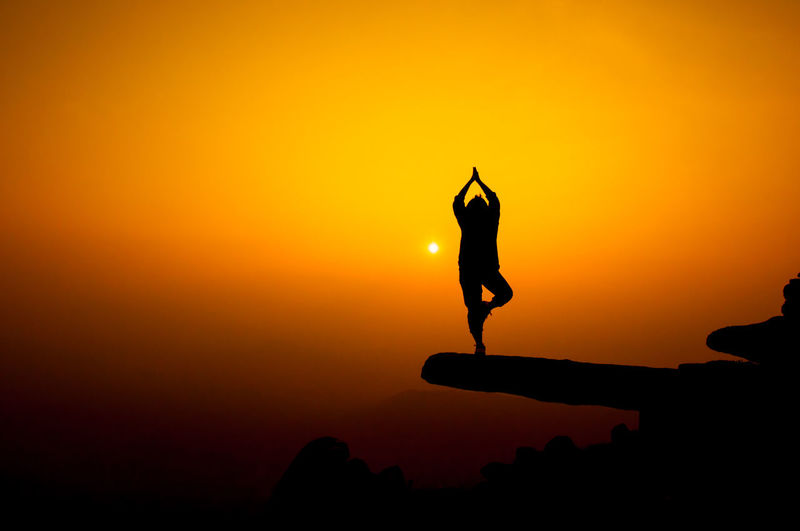 Silhouette person doing yoga on cliff against sky during sunset