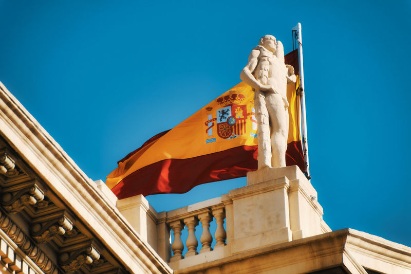 Low angle view of historical building and spanish national flag against blue sky, mnac, barcelona 