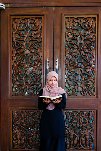 Woman wearing hijab while reading holy book against wood door