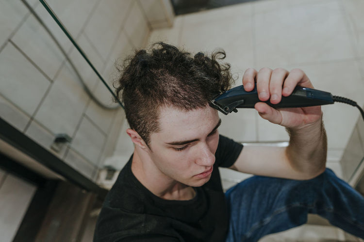 Sad young man with electric razor shaving his own hair in bathroom