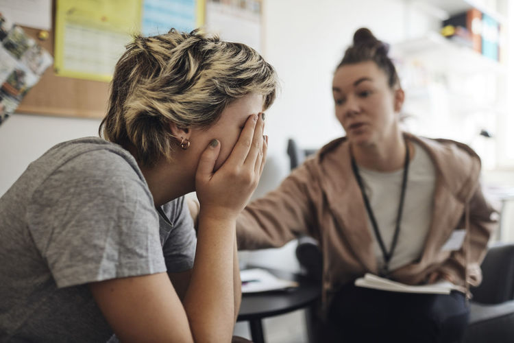 Non-binary counselor consoling female student covering face with hand in school office