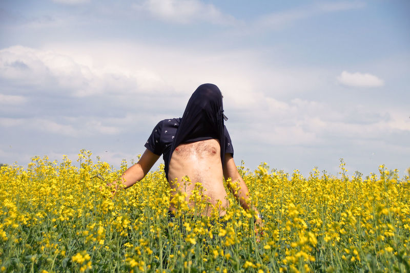 Man covering face with t-shirt while standing amidst yellow flowers on oilseed rape field
