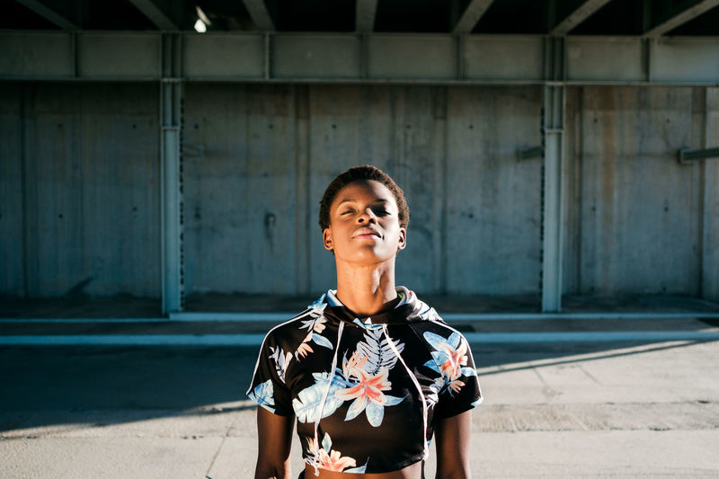 High angle of charismatic confident african american female athlete in flowered sportswear with closed eyes while standing alone on street in sunbeams against concrete wall in city