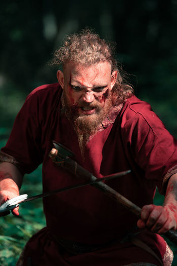 Furious male warrior shouting aggressively during attack with sword and axe at viking reenactment in forest
