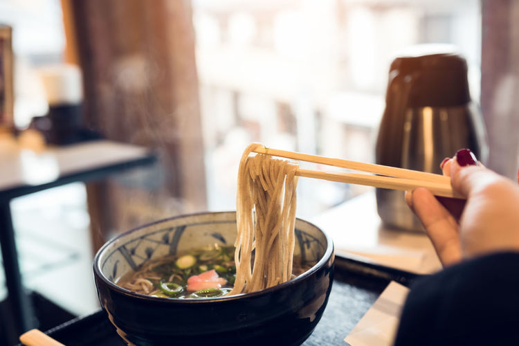 Cropped hand holding noodles with chopsticks in bowl on restaurant table