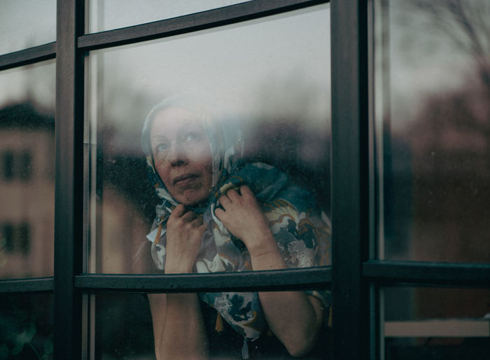 Sad mature woman with scraft on head standing behind dirty window glass