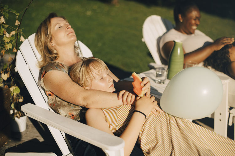 Mother relaxing with daughter in backyard on deck chair during sunny day