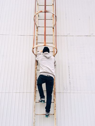 Rear view full length of man climbing on rusty ladder by white wall