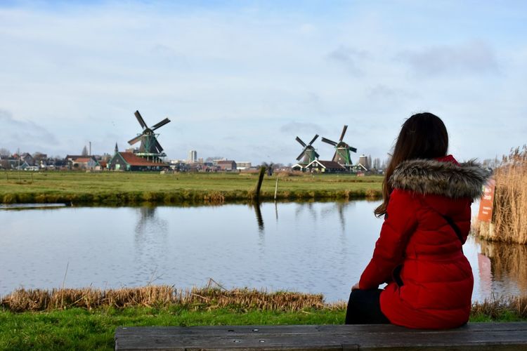 Woman sitting on bench by lake against windmills