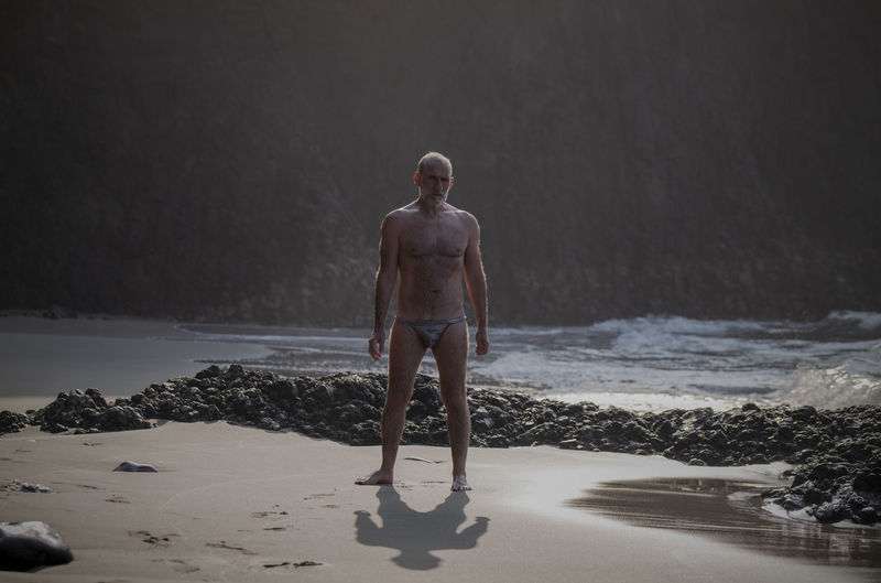 Full length of shirtless man  with swimwear standing on beach 