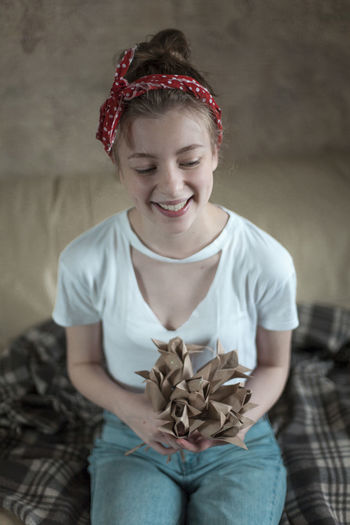 Cheerful young woman holding paper flowers while sitting on bed