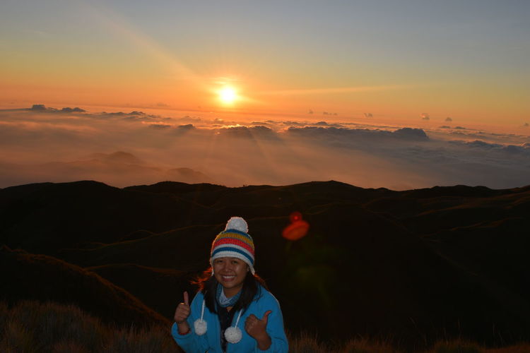 Smiling young woman with thumbs up on mountain at sunset