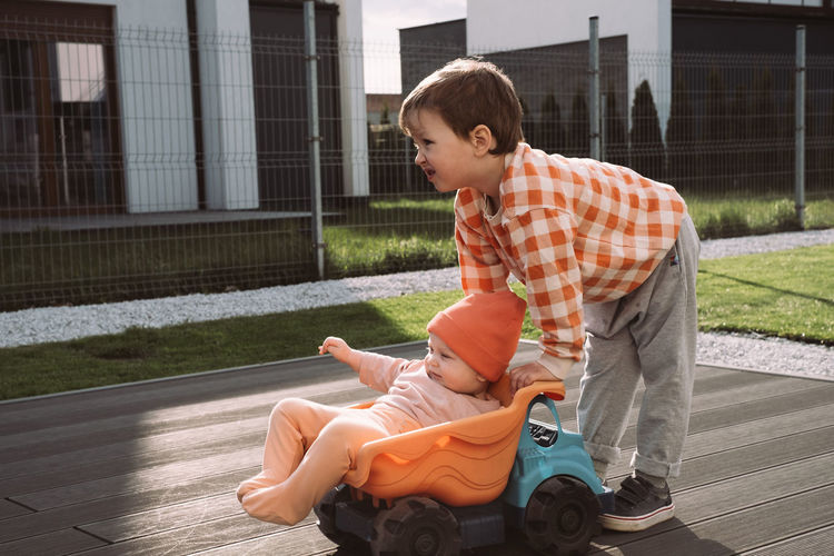 Brother giving a ride to baby sister in big toy car outdoors. children having fun near house. boy