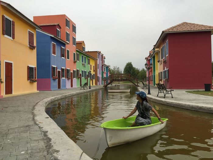 Girl sitting in boat moored at canal amidst residential buildings against sky