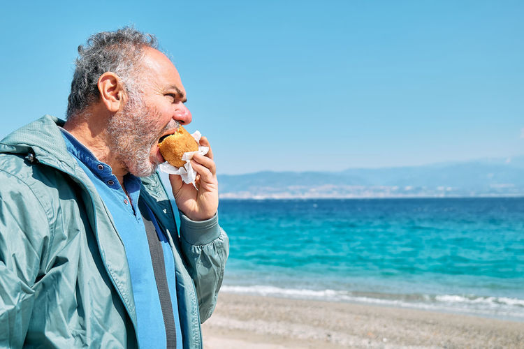 Bearded mature man at spring seaside eating hot palatable arancina