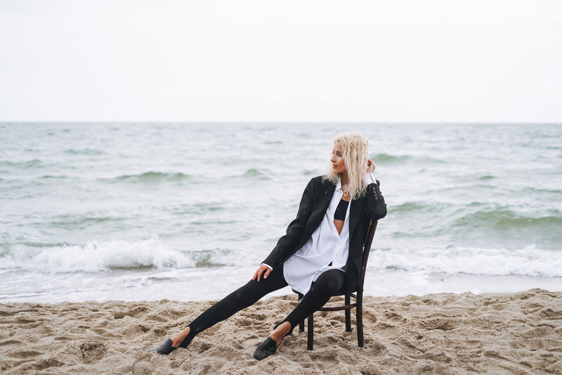 Portrait of elegant blonde woman in black suit on vintage wooden chair by sea in a storm