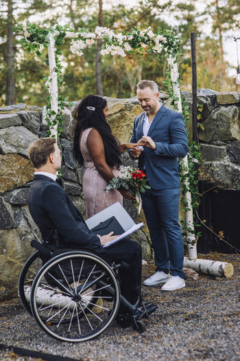 Smiling groom and bride exchanging ring by minister in wheelchair with disability at wedding ceremony