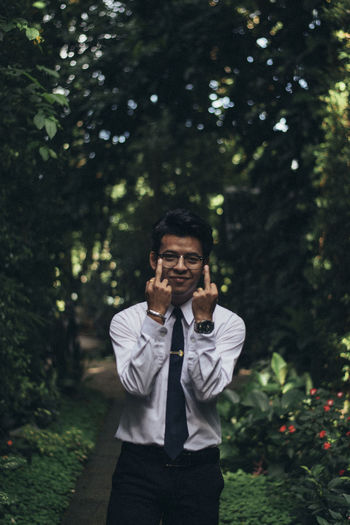 Portrait of smiling young man gesturing while standing on footpath against trees