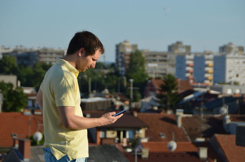 Man using phone by buildings in city against clear sky