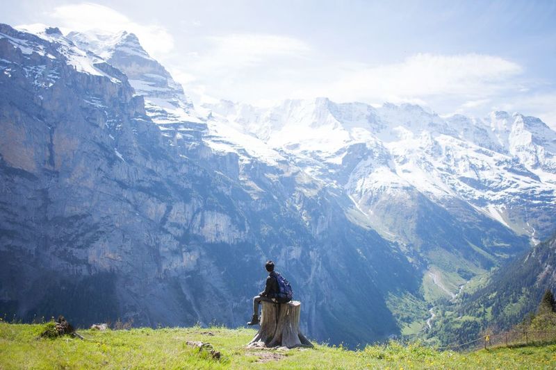 Rear view of young hiker sitting on tree stump in front of snowcapped mountains