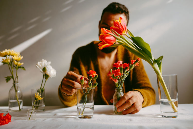 Anonymous male florist sitting at table with carnations and tulips in glassware