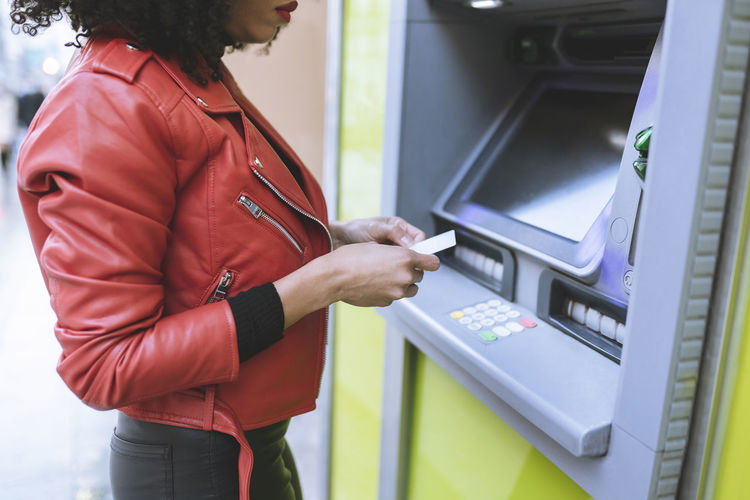 Anonymous african american female standing near atm and inserting credit card while smiling widely and looking away