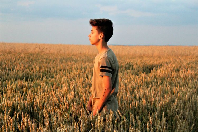 Side view of teenage boy standing amidst crops in field against sky during sunset