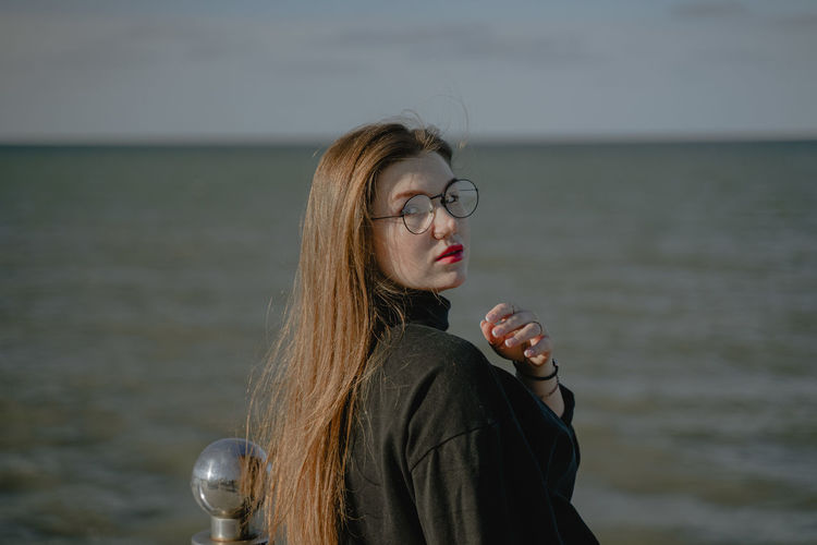 A girl with long red hair and round eyeglasses looks back at the seashore, with red lipstick 
