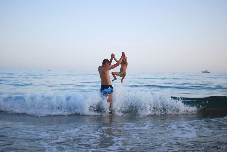 Full length of father and son jumping in sea against clear sky