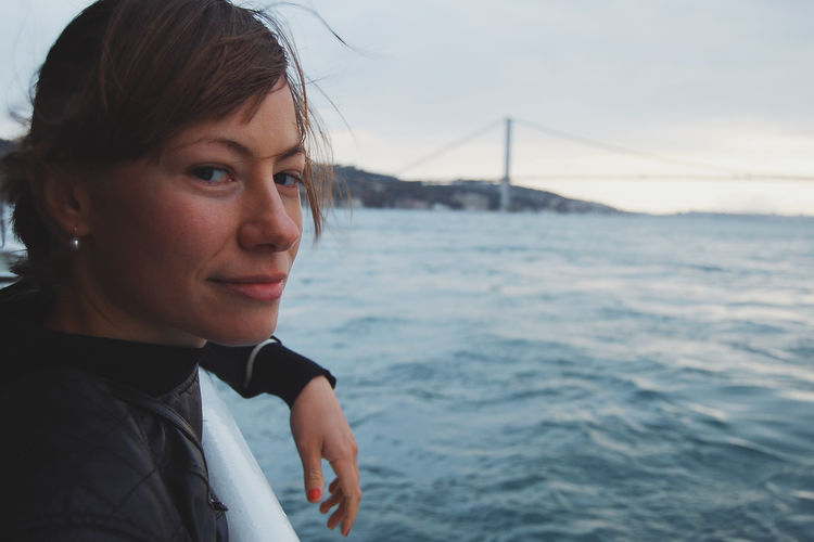 Close-up portrait of young woman traveling on ferry at sea against bridge