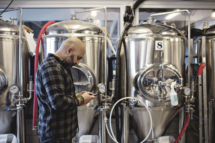 Side view of manager using mobile phone while standing by storage tanks at brewery