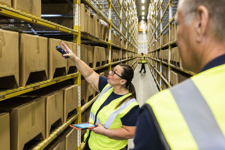 Female worker holding digital tablet while pointing at cardboard box on rack to coworker in distribution warehouse
