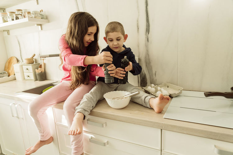 Girl with brother holding shakers over bowl sitting on kitchen counter at home