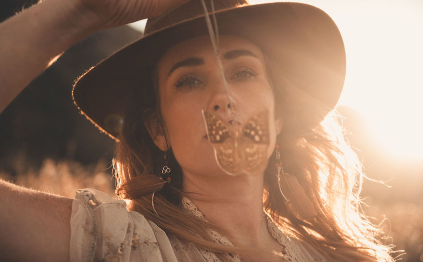Portrait of beautiful woman wearing hat while holding pendant during sunset