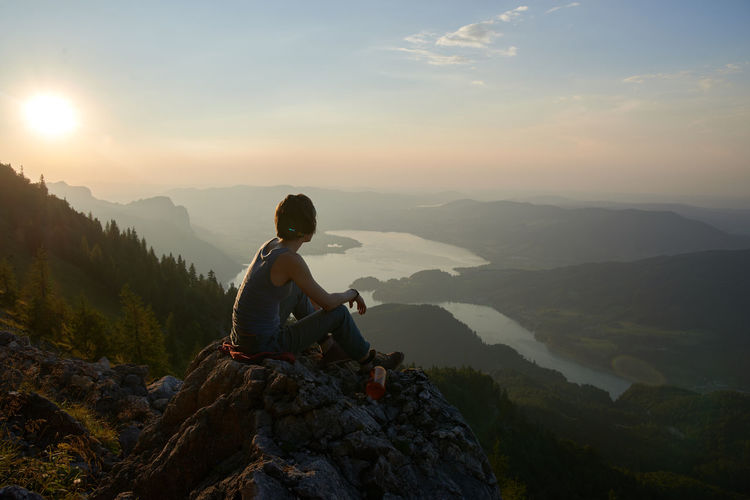 Hiker sitting on cliff against sky during sunset