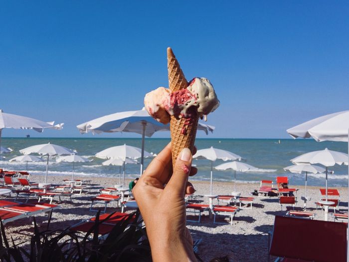 Cropped hand holding ice cream cone at beach against clear blue sky