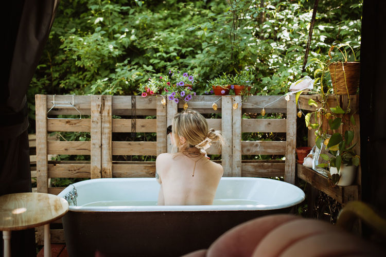 Back view of faceless topless thoughtful female sitting in bathtub near wooden fence on patio during skin care routine on summer day