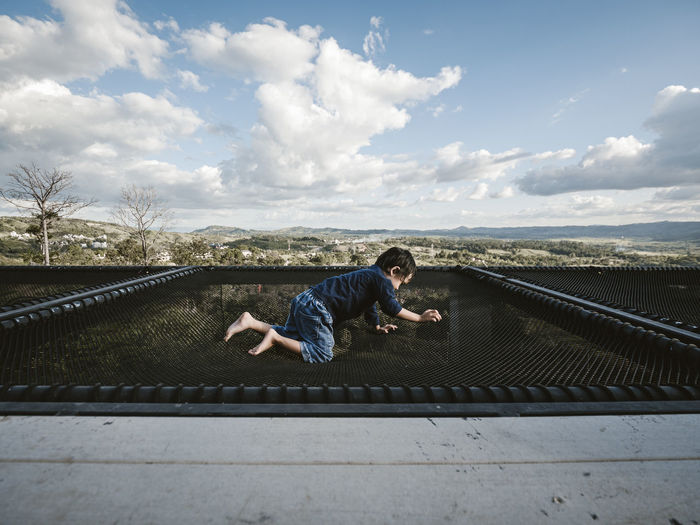 Side view of boy crawling on netting against sky