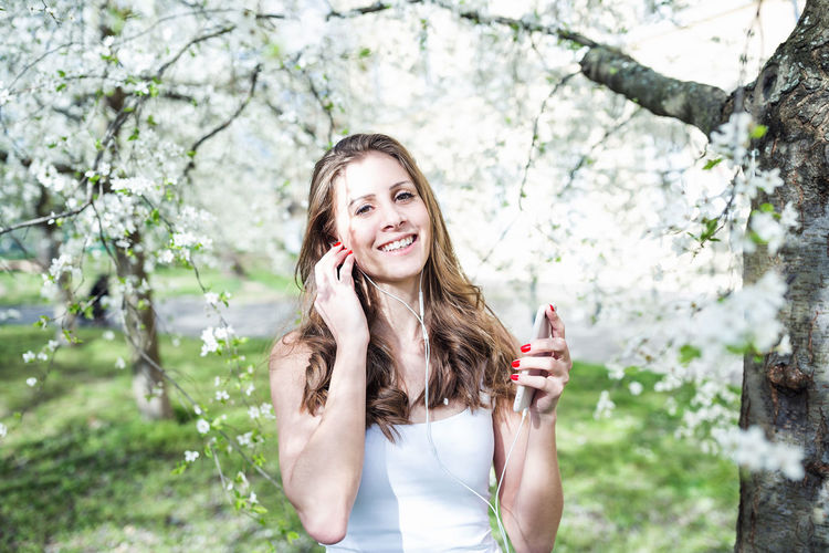 Young ukrainian woman listening to music or podcast on phone under blooming trees in spring park