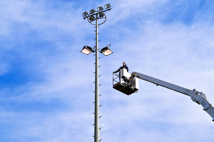 Low angle view of man on cherry picker by floodlight against sky