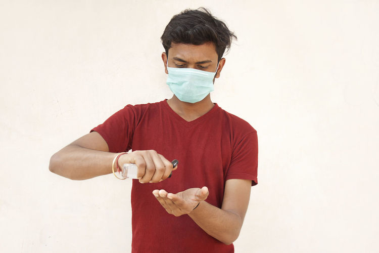 Young man wearing flu mask using hand sanitizer standing against white background