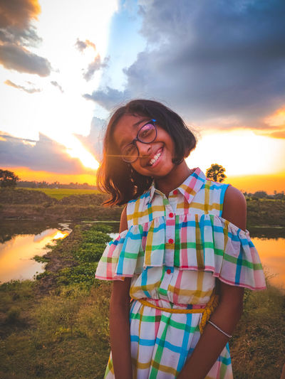 Portrait of smiling young woman standing on land against sky during sunset