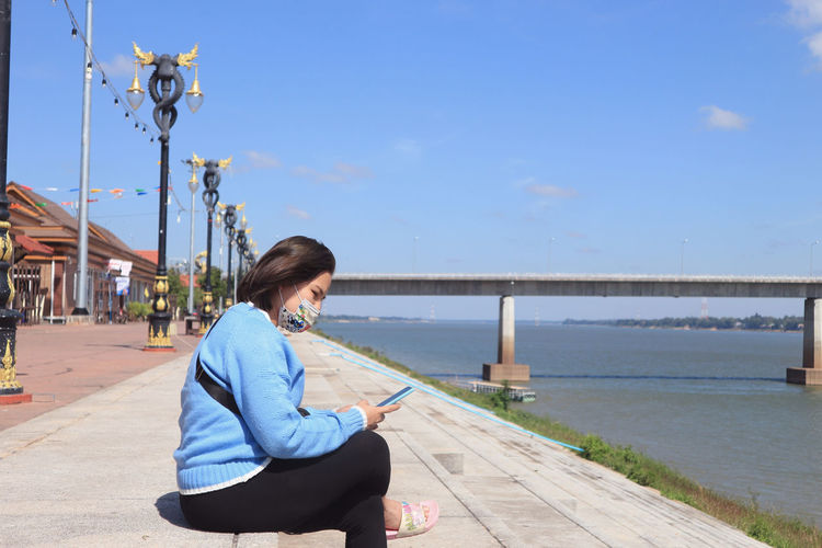 A woman sitting holding a smartphone at bridge across the mekong river that connects mukdahan.