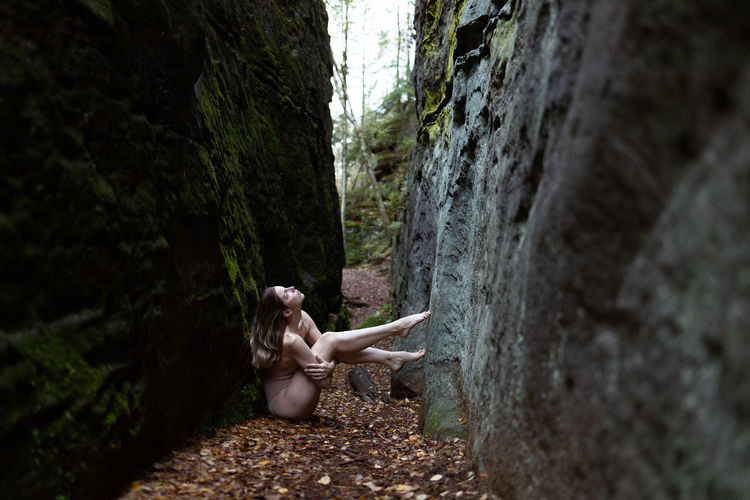 Side view of naked woman sitting amidst rock in forest