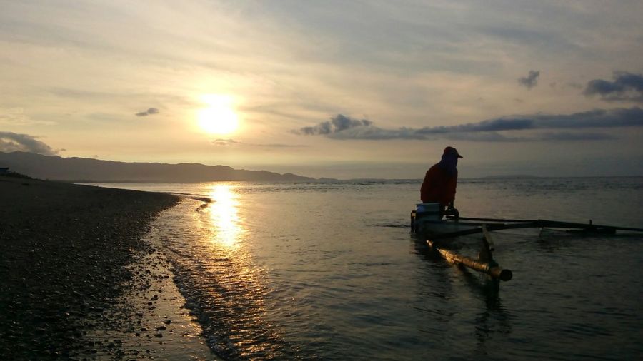 Rear view of fisherman in sea against sky during sunset